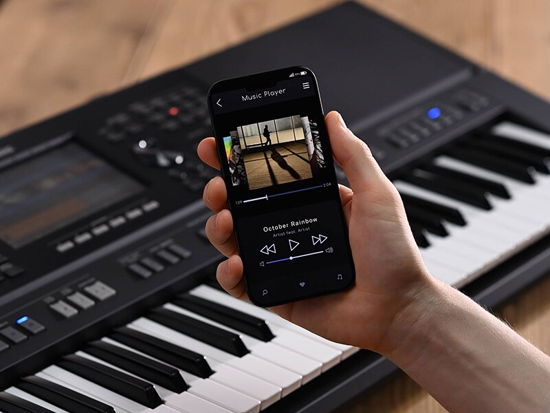 Yamaha Arranjador Workstation PSR-SX920 e uma mão segurando um smartphone com a tela de um player de música