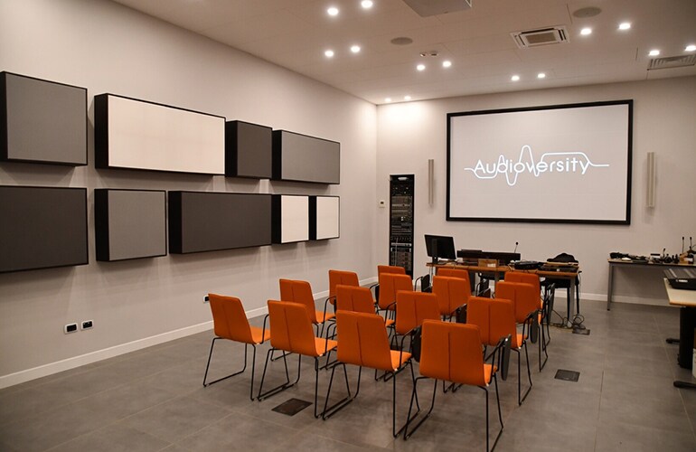 Centro de treinamento de Lesmo, com cadeiras laranja, tela de apresentação e equipamentos de áudio montados na parede.